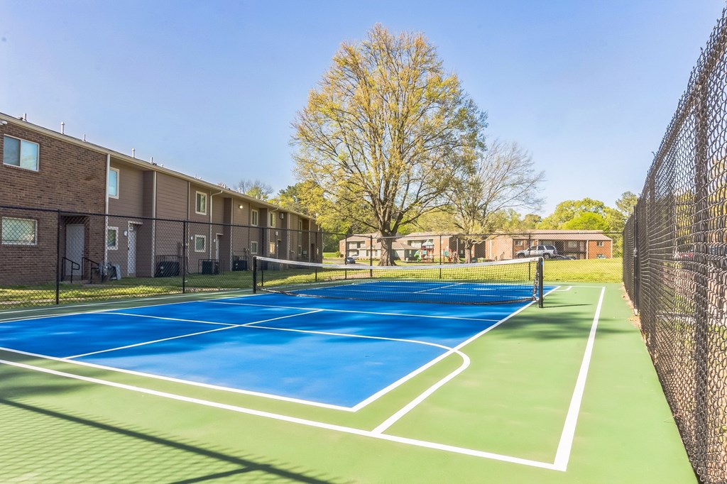 A tennis court is surrounded by a chain link fence and apartment buildings.