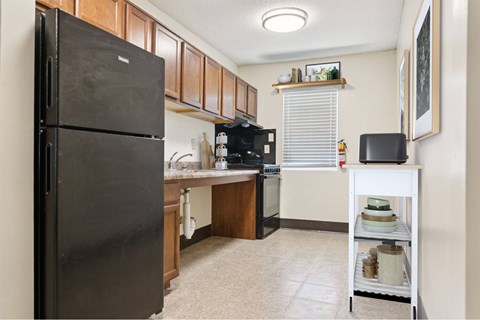 A black refrigerator stands in a kitchen with wooden cabinets and a white shelf.