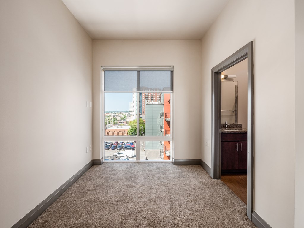 A room with a carpet floor and a view of a parking lot through a window.