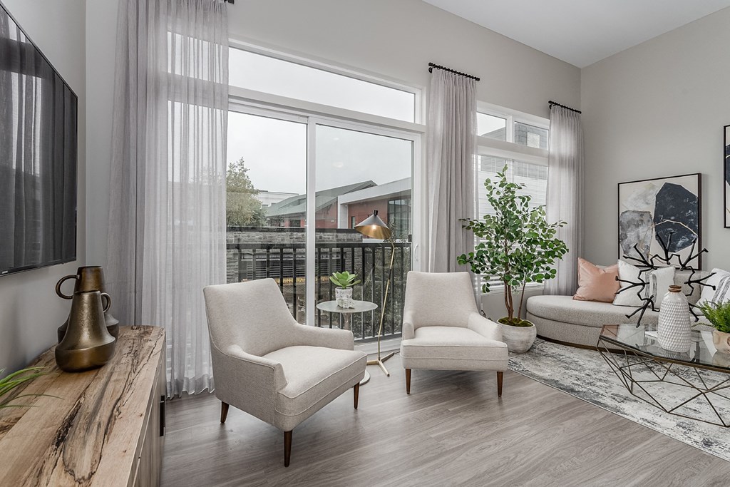 2-Bedroom Apartments in Liberty Township, OH - Liberty Center - Furnished Living Room with Vinyl Wood Plank Flooring, Area Rug, Sofa, Coffee Table, Armchairs, Houseplant, Storage Cabinet, Wall-Mounted TV, Large Window, and Sliding Glass Door with Access to Balcony.