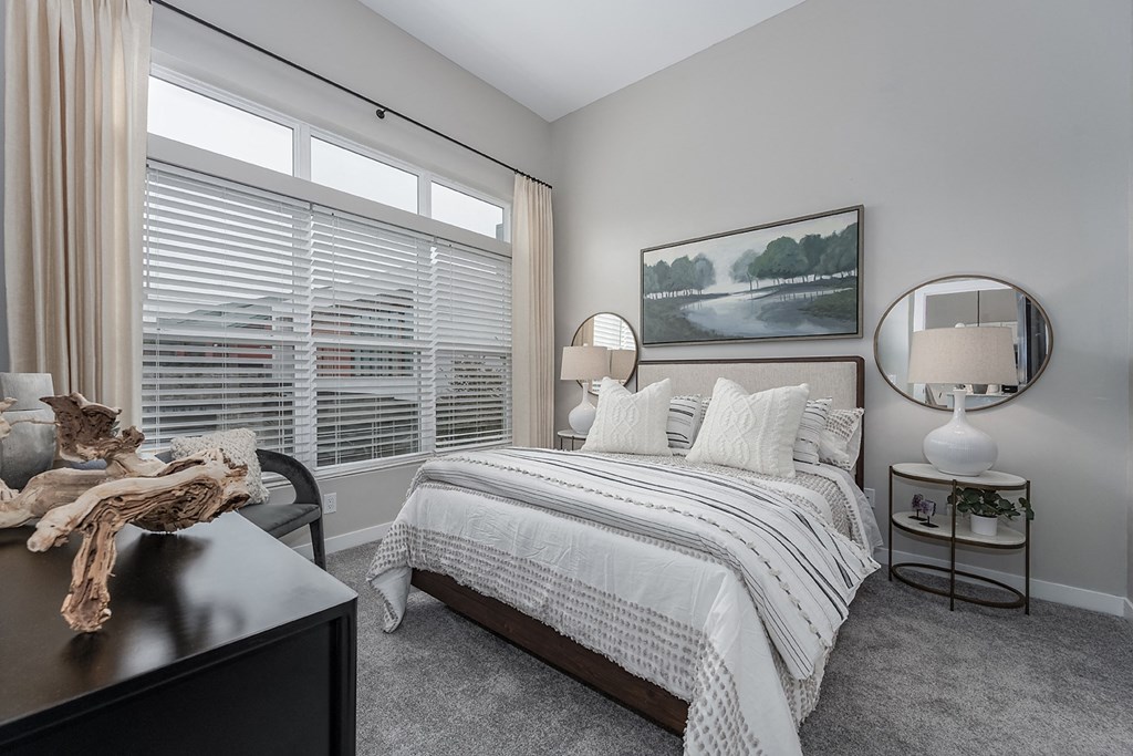 One-bedroom Apartments in West Chester OH - Liberty Center - Furnished Bedroom with Grey Shaw Carpeting, Round Coffee Tables with Lamps, Circular FRamed Mirrors, Chair, Wooden Dresser, Window