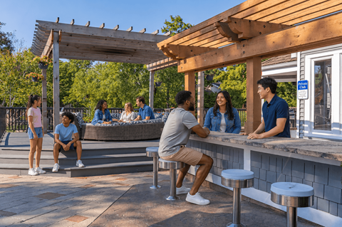 A group of people are sitting and standing around a picnic table under a wooden pergola.