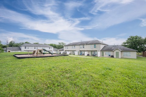 A large grassy field with a playground and houses in the background.