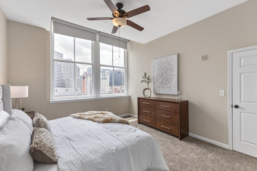 A bedroom with a bed, a ceiling fan, and a window overlooking a cityscape.