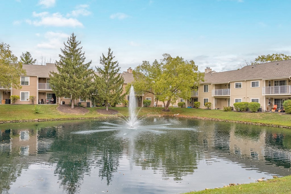 A fountain in the middle of a pond in front of apartment buildings.