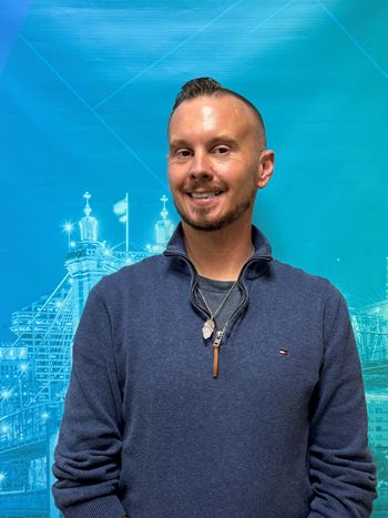 A man with a necklace and a blue shirt is standing in front of a blue background with a building.