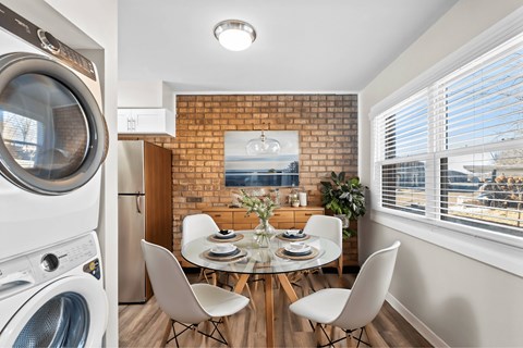 A modern laundry room with a washing machine and dryer on the left and a dining area with a table and chairs in the middle at Fountain Parc Apartments, Indianapolis, 46220