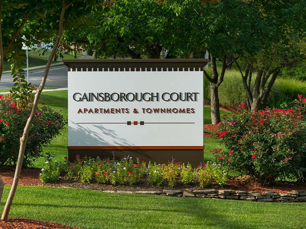 A sign for Gainsborough Court Apartments & Townhomes is surrounded by flowers and trees at Gainsborough Court Apartments, Fairfax, 22030