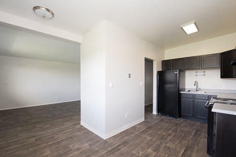 an empty living room with a kitchen and a black refrigerator