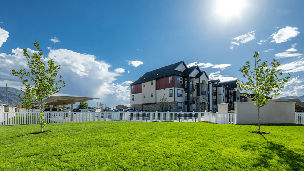 A white picket fence surrounds a green lawn with two trees and a large building in the background.