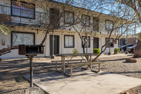 A wooden picnic table is in front of a building with a tree in front of it.