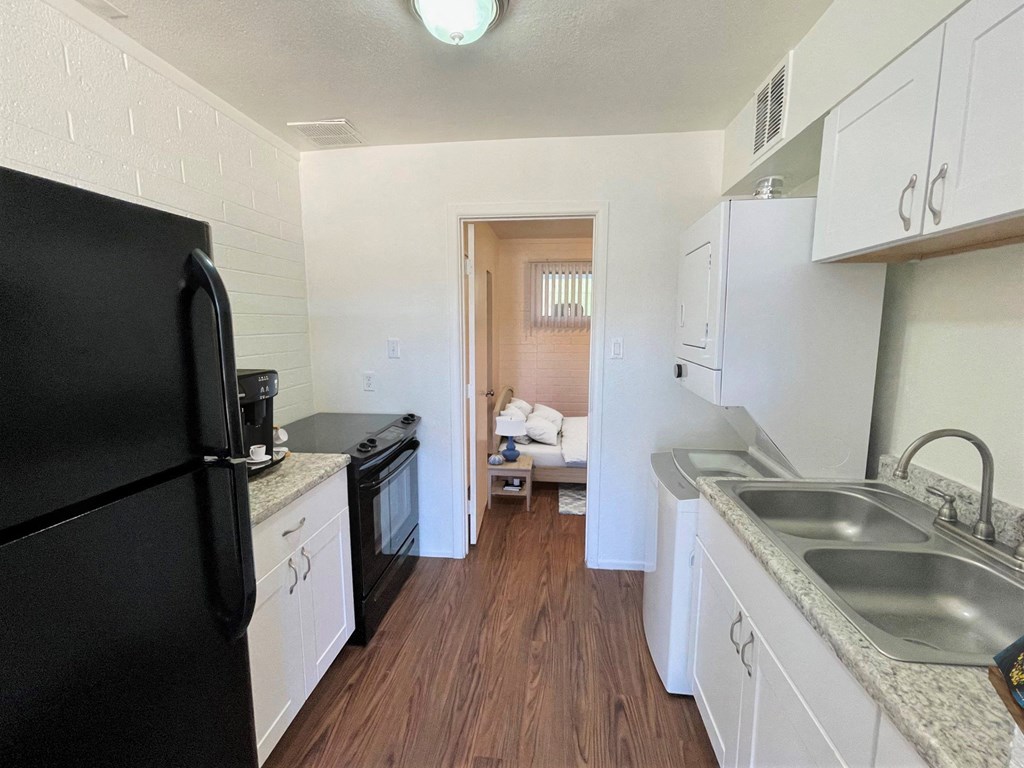 A black refrigerator stands in a kitchen with white cabinets and a wooden floor.
