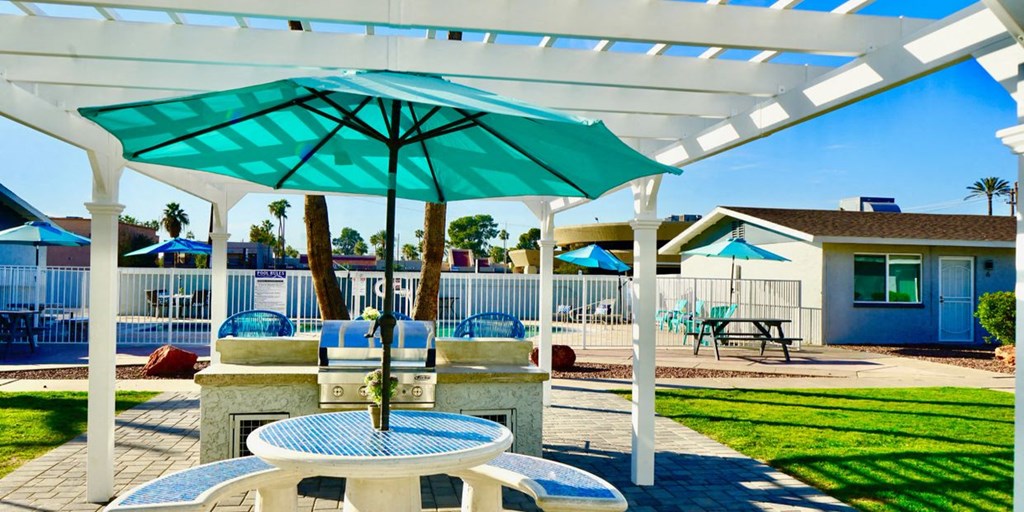 A white pergola with a blue umbrella is over a table with chairs.