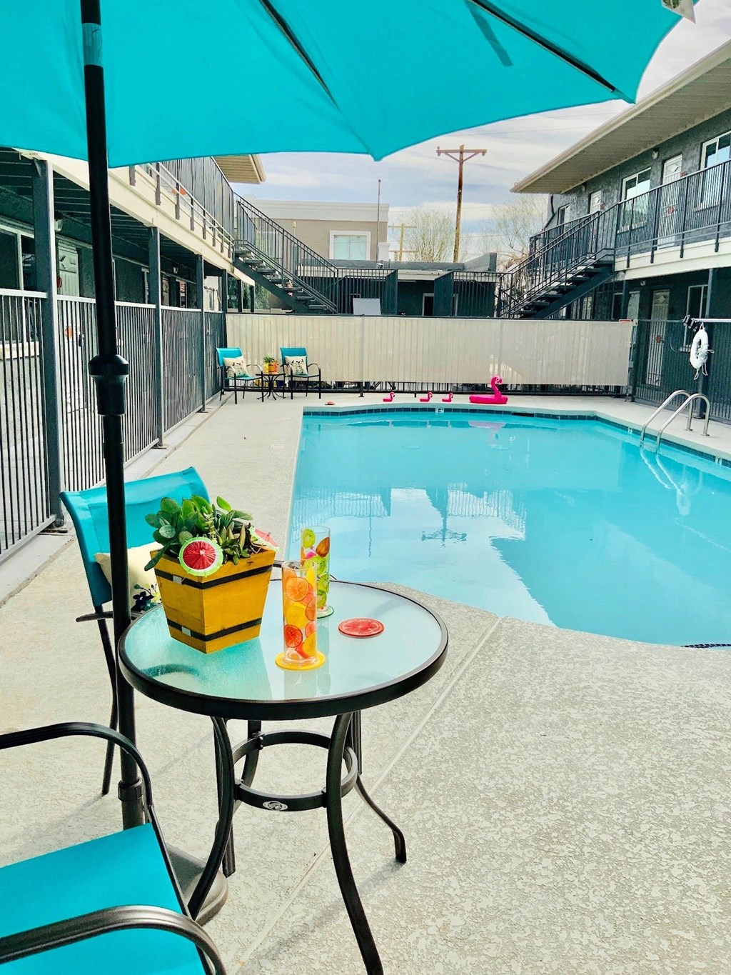 A table with a yellow container and a glass of orange juice is on a patio next to a pool.