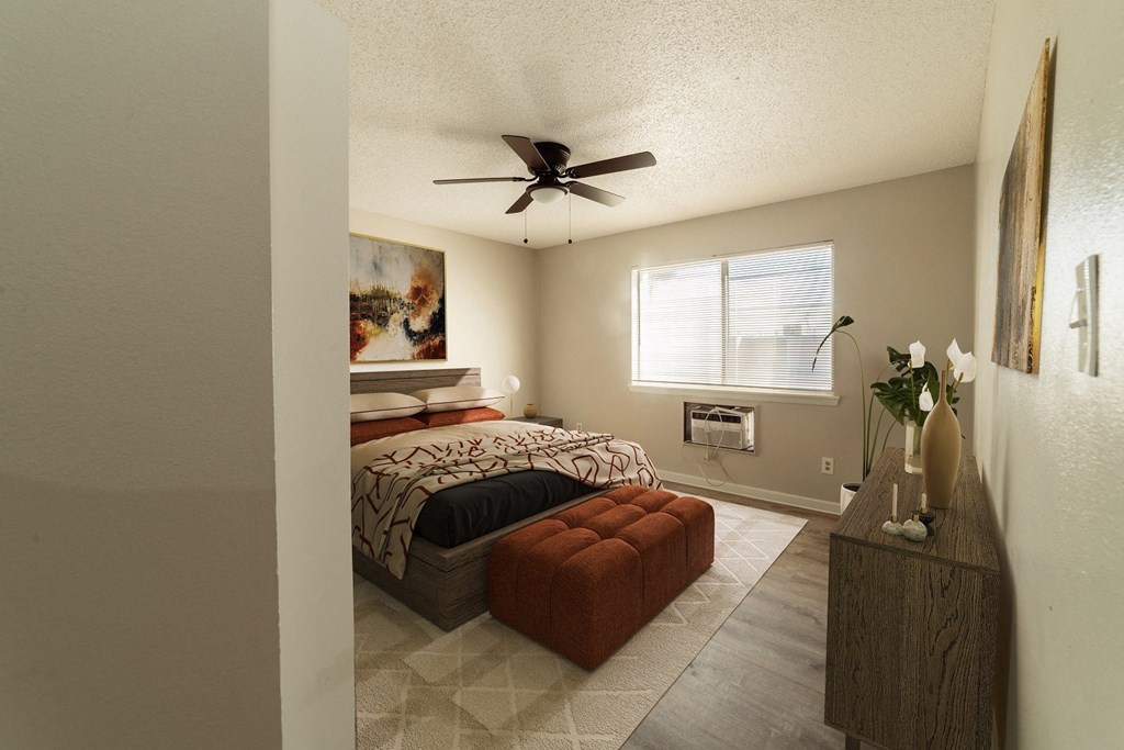 A bedroom with a bed, a red ottoman, and a ceiling fan.