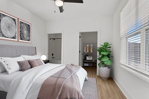 Staged apartment bedroom at Windsor Sugarloaf Heights 5805 Sugarloaf Parkway Lawrenceville GA 30043 featuring a bed with upholstered headboard wood style flooring ceiling fan natural light and open access to the bathroom and walk in closet