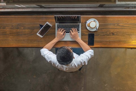 A man in a white shirt is typing on a laptop with a smartphone and a cup of coffee on the table.