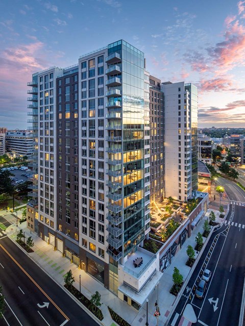 A modern multi-story building with glass windows is illuminated from within and surrounded by a well-lit street.