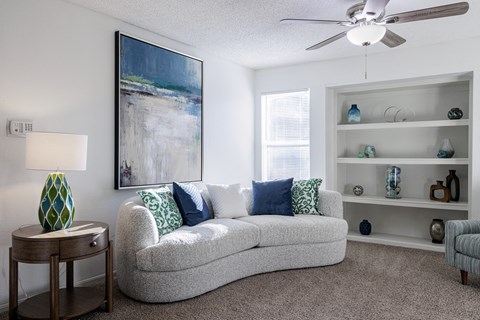 A living room with a grey couch, a painting on the wall, a side table with a lamp, and a shelf with decorative items.