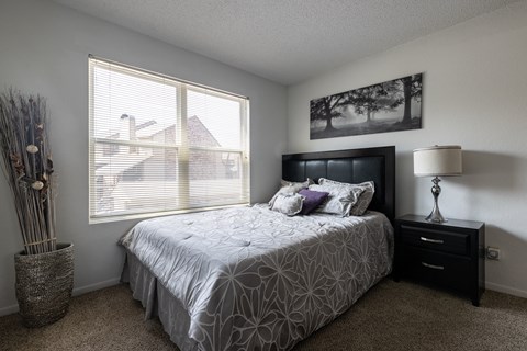 A bedroom with a large bed, a nightstand with a lamp, and a window with blinds.