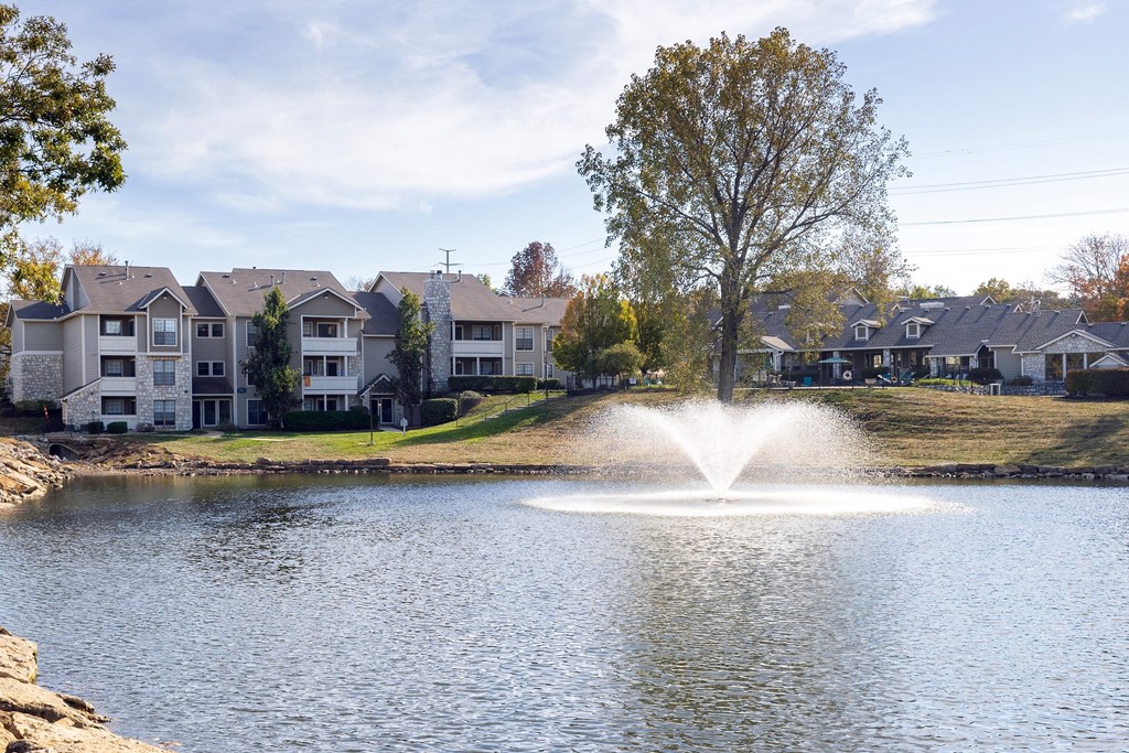 Serene lake with a fountain at the Three Lakes apartment complex, with the apartment buildings in the background and greenery all around.