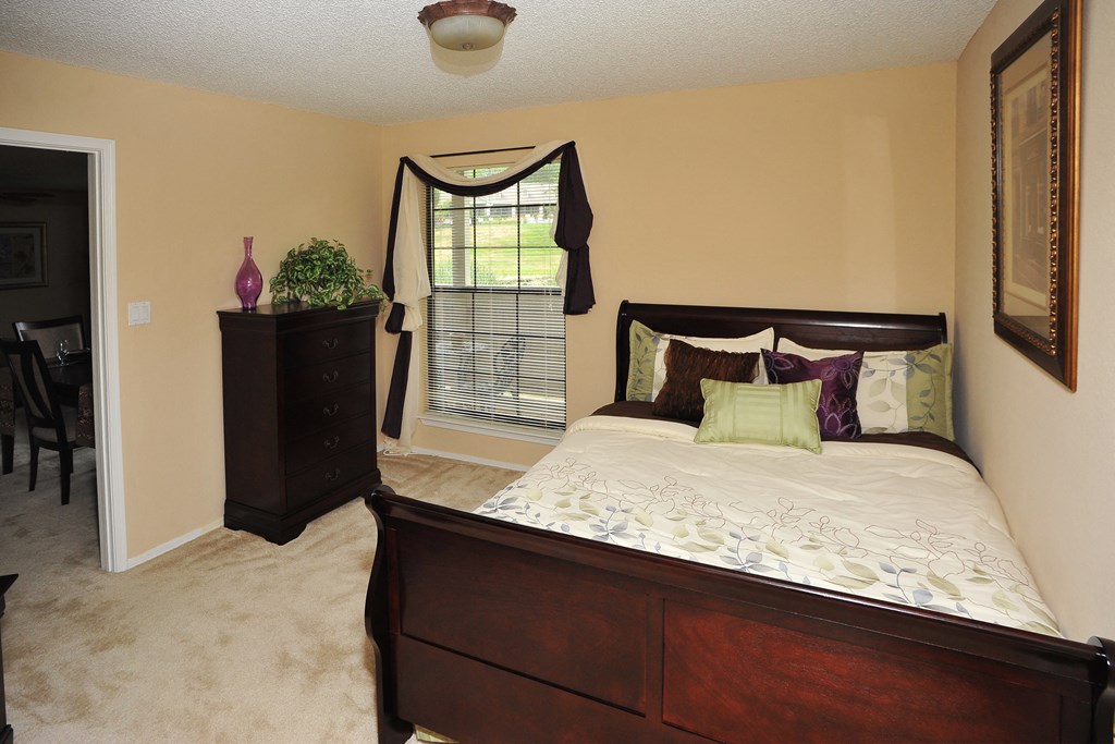 Cozy bedroom with a dark wood bed and dresser, beige walls, white bedding, a window bringing in natural light, and carpeted floors.