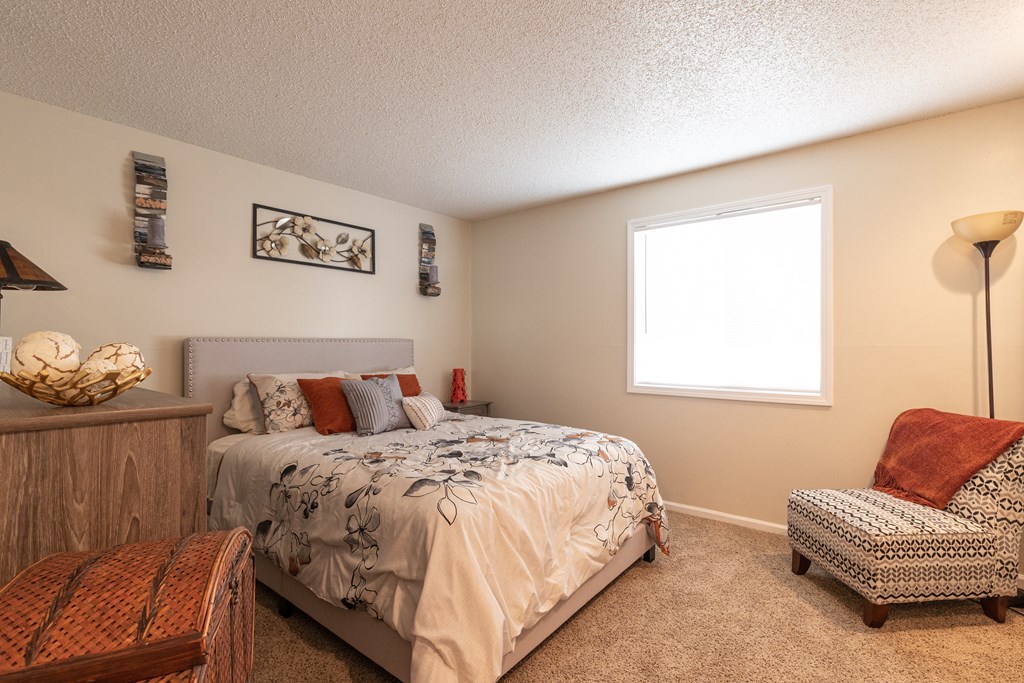 A spacious bedroom featuring a beige padded headboard, floral black-stitch bedding, a natural wood dresser on the left, a cozy orange lounge chair on the right, white walls, and a window offering a scenic view.
