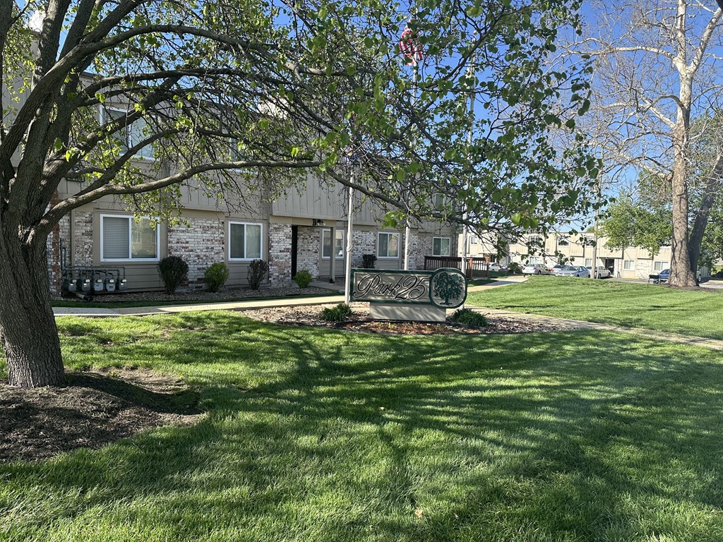 Exterior view of Park 25 Apartments, featuring a house with a green sign in the grass, a tree, and lush green grass surrounding the apartment building.