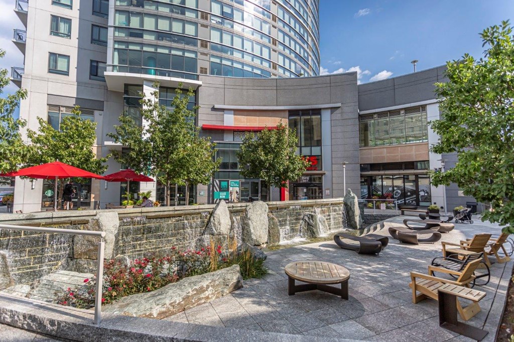 A modern building with a glass facade is surrounded by a stone wall with a garden and seating area in front.