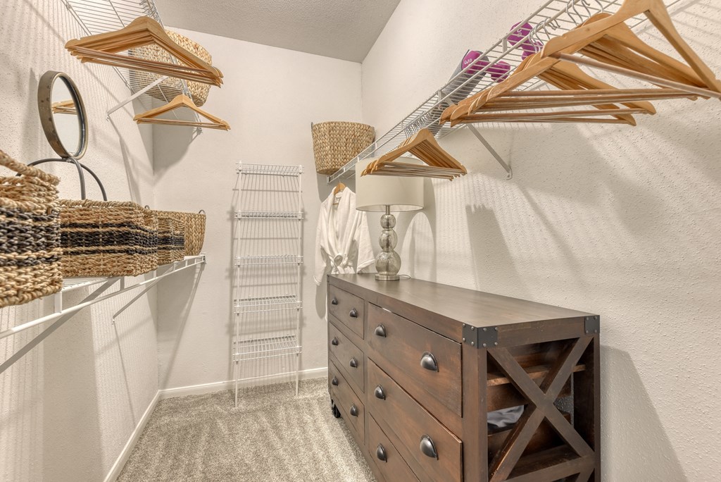 a walk in closet with white walls and a wooden dresser with a lamp on it