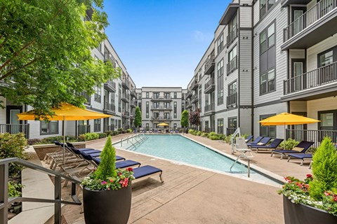 A pool area with sun loungers and umbrellas in front of apartment buildings.