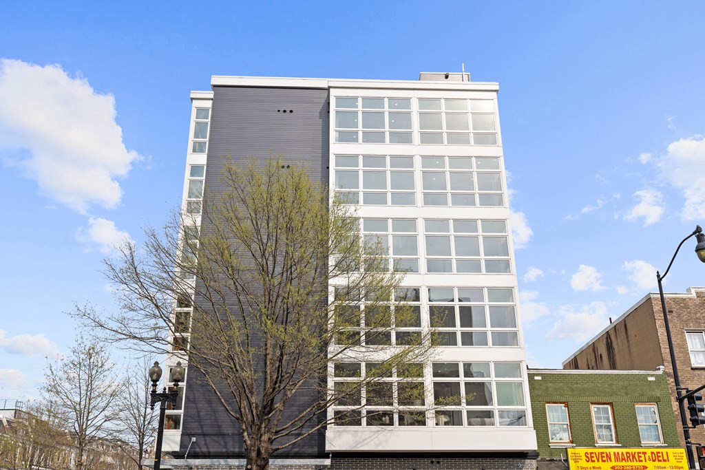 A tall building with a grey top floor stands next to a tree and a smaller building.