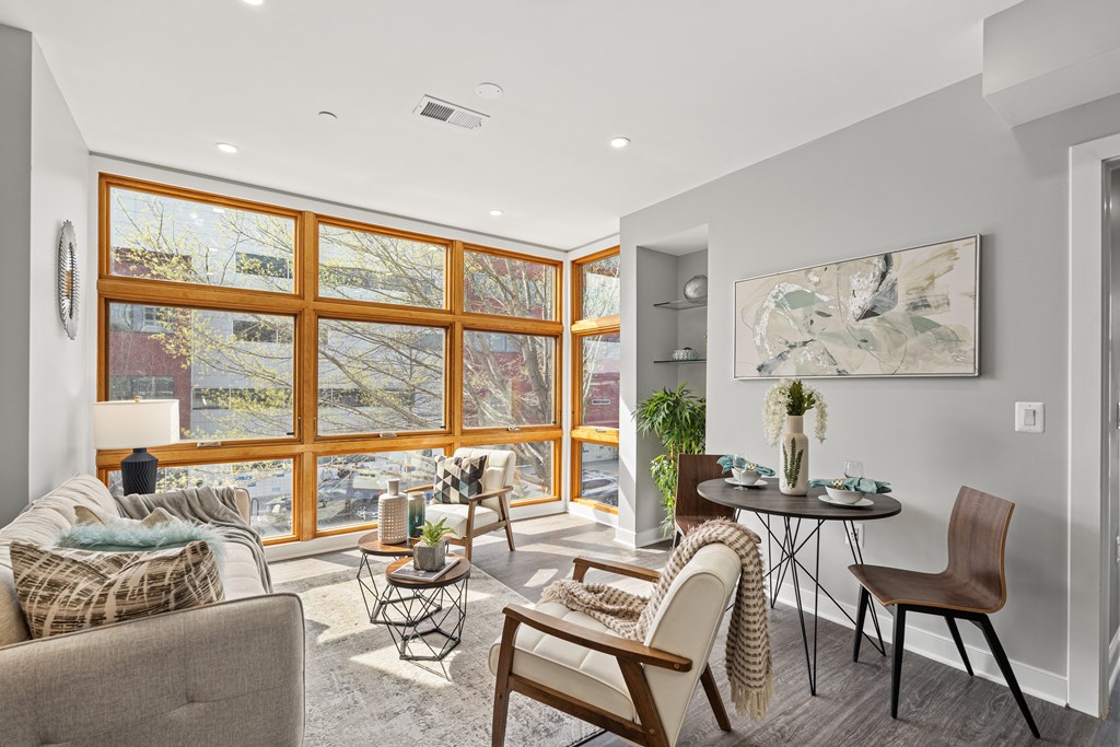 A living room with a grey couch, a wooden chair, a small table with a vase on it, and a large window with a view of trees.
