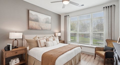 A well-lit bedroom with a large bed, a painting on the wall, and a window with blinds at Vintage Raleigh West Apartments, Raleigh, North Carolina