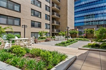 A patio with chairs and tables is surrounded by a building and a green area.