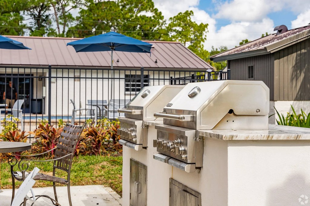 A grill is on a table outside with a blue umbrella and chairs.