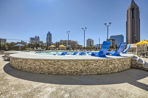 A round stone pool with blue loungers and umbrellas in the foreground with a city skyline in the background.