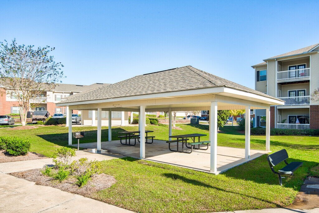 A covered picnic area with benches and a table is surrounded by a grassy area.