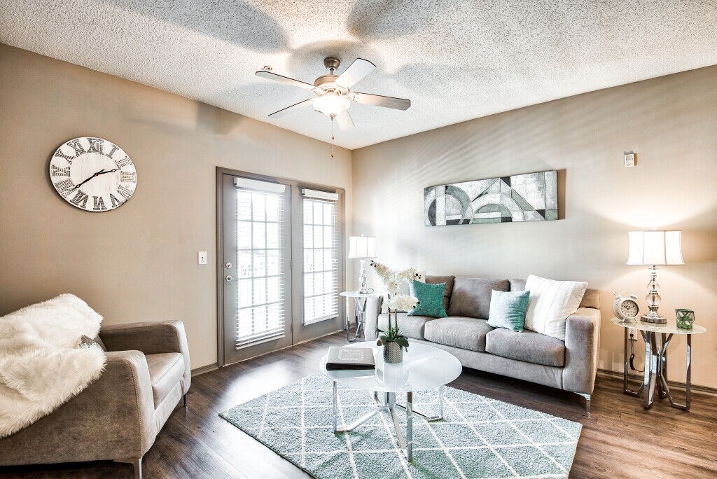A living room with a grey couch, a white chair, a clock on the wall, and a ceiling fan.