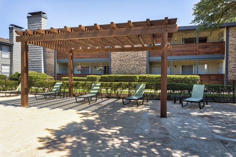 A patio with a pergola and chairs is surrounded by apartment buildings.