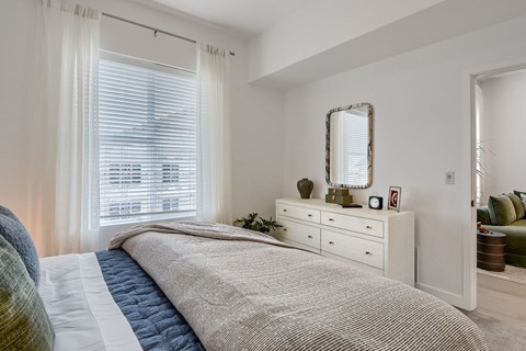 A bedroom with a large bed, a dresser with a mirror, and a window with white curtains.