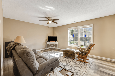 A living room with a grey couch, a wooden chair, a white table, and a ceiling fan.