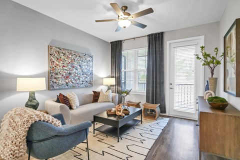 A living room with a grey armchair, a white sofa, a coffee table, a ceiling fan, and a large abstract painting on the wall.