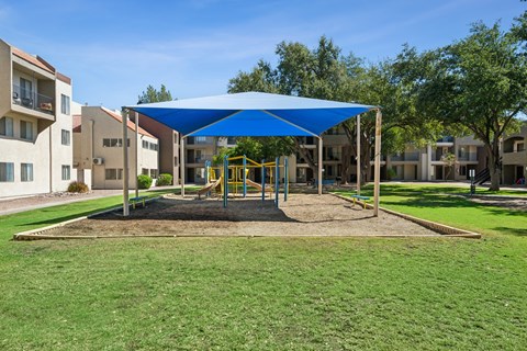 A playground with a blue canopy and yellow equipment is surrounded by apartment buildings.