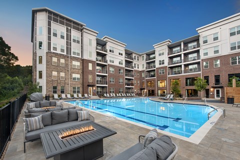 A large outdoor pool with a fire pit and lounge chairs is surrounded by apartment buildings.