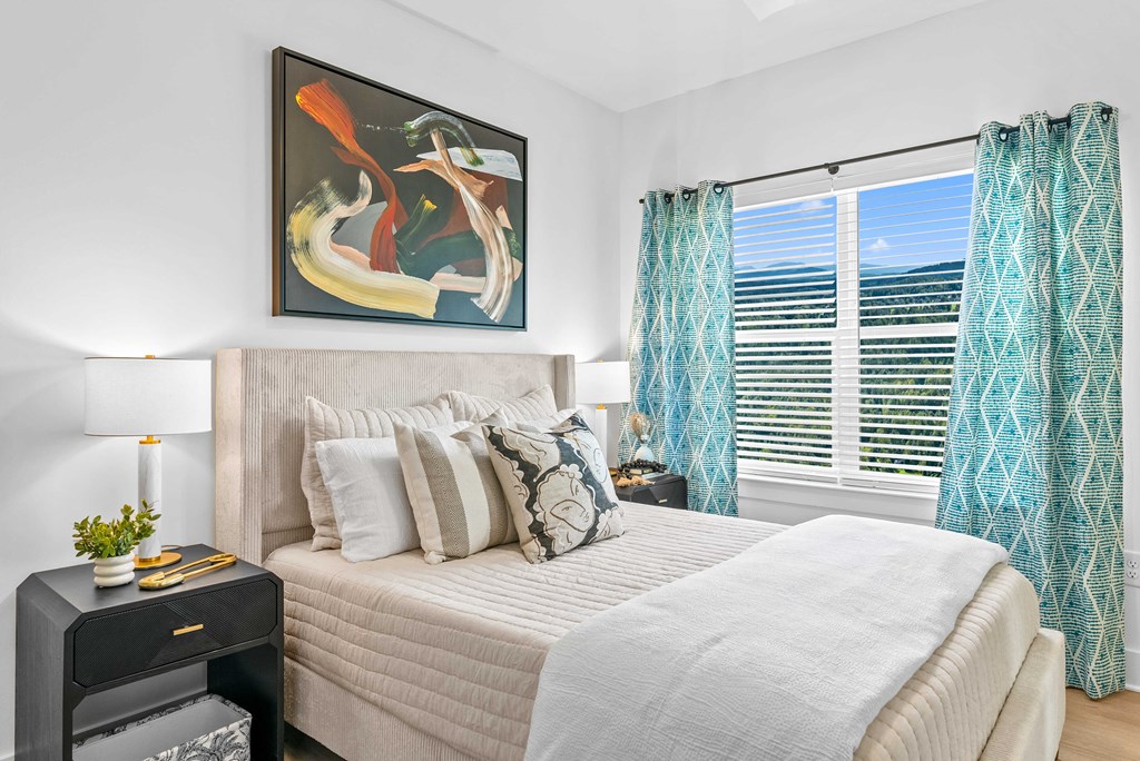 A bedroom with a large bed, a painting on the wall, and a window with blue curtains.