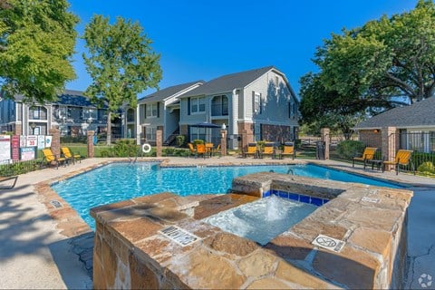 A pool with a hot tub in front of a building with a fence and trees.