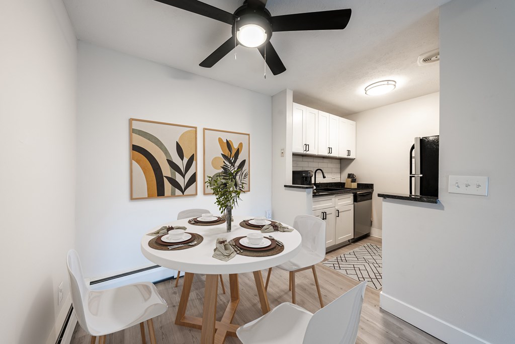A dining room with a white table and chairs, a ceiling fan, and a kitchen area in the background.