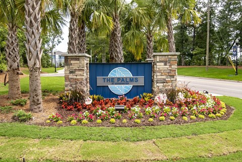 A sign that says "The Palms" is surrounded by flowers and palm trees.