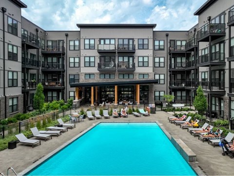 A large swimming pool is surrounded by lounge chairs in front of apartment buildings.